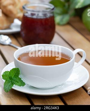 Tazza di tè con foglie di menta fresca e croissant e barattolo di marmellata sullo sfondo su un tavolo da patio in legno Foto Stock