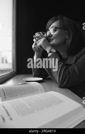 Ritratto in bianco e nero di una donna al bar. Donna in occhiali con libro aperto, bianco e nero. Donna matura che sogna in un bar. Concetto di autostima. Foto Stock