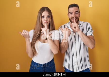 Giovane coppia in piedi su sfondo giallo che chiede di stare silenziosa con un dito sulle labbra che punta con la mano di lato. silenzio e concetto segreto. Foto Stock