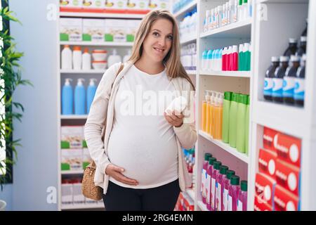 Giovane donna incinta cliente che tocca la pancia tenendo pillole scaffalatura bottiglia in farmacia Foto Stock