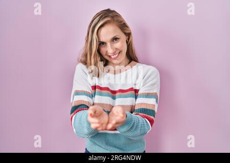 Giovane donna bionda in piedi su sfondo rosa sorridente con mani palme insieme ricevendo o dando gesto. Tenere e protezione Foto Stock