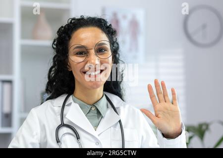 Video consultazione online, medico femminile in cappotto medico bianco che parla su videochiamata sorridendo e guardando la macchina fotografica, donna latino americana con stetoscopio che saluta gioiosamente gesto di saluto. Foto Stock