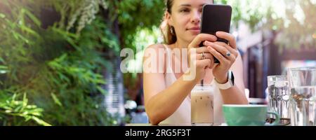 Smartphone nelle mani di una giovane donna seduta in una caffetteria sul marciapiede in estate sullo sfondo. Foto Stock