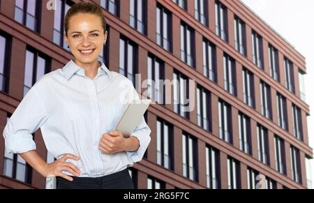 Giovane agente immobiliare d'affari in piedi di fronte al business center in centro, con un tablet digitale in mano e sorridente. Foto Stock