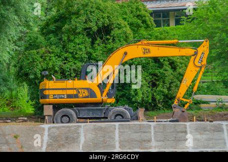 Vista frontale dell'escavatore cingolato per scavo in cantiere di demolizione in controluce. un escavatore sullo sfondo verde in un parco cittadino Foto Stock