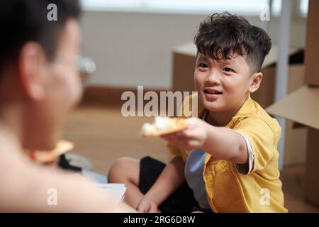 Un ragazzo che condivide una fetta di pizza con papà seduto sul pavimento in un nuovo appartamento Foto Stock