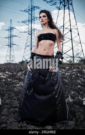 Moda informale: La bellissima ragazza goth sottile vestita con gonna e guanti in pelle nera. Ritratto all'aperto nel campo vicino a piloni e linee elettriche Foto Stock