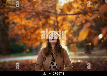 Ciao november. una donna di mezza età sorridente e alla moda, con cappotto beige e cappello arancione, lancia foglie autunnali all'esterno del parco cittadino in autunno. Foto Stock