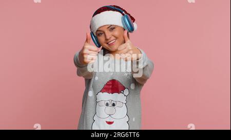 Felice giovane donna natalizia in cuffia ascoltando musica e ballando discoteca, divertendosi a giocare con le mani gesticolate espressive e rilassandosi durante la festa. Adorabile ragazza isolata su sfondo rosa studio Foto Stock