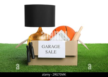 Articoli diversi nella scatola con cartello Garage sale su erba verde su sfondo bianco Foto Stock