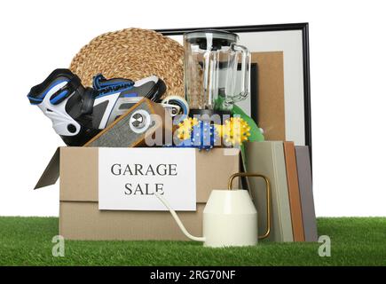 Scatola con cartello Garage sale e cose diverse su erba verde su sfondo bianco Foto Stock