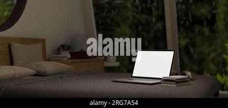 Immagine ravvicinata di un mockup con schermo bianco di un notebook, libri e cuffie su un letto in una camera da letto durante una notte di pioggia. Concetto di spazio di lavoro domestico. rendering 3d, 3d il Foto Stock