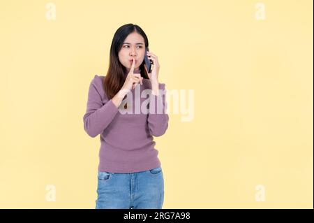 Una bella donna asiatica sta scherzando o raccontando un segreto alla sua amica al telefono, in piedi su uno sfondo giallo isolato. Foto Stock