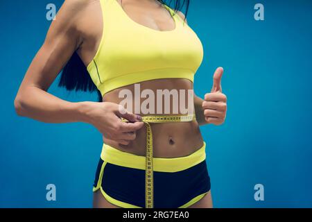 Crop colpo di donna fitness in sport outfit girovita misura metrica con nastro e mostrando il pollice verso l'alto. Buoni risultati di esercizio in palestra. Forma perfetta Foto Stock