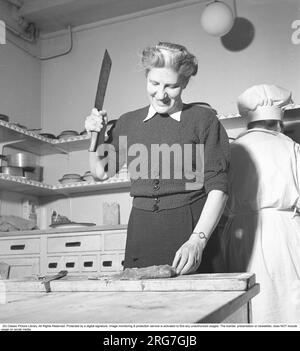 Donna felice in cucina nel 1948. Una donna in una cucina del ristorante si alza e taglia la carne a pezzi con un coltello più grande. Ha messo il pezzo di carne su un tagliere e ha alzato il coltello da macellaio per iniziare. Svezia 1948. Kristoffersson rif. AF26-4 Foto Stock