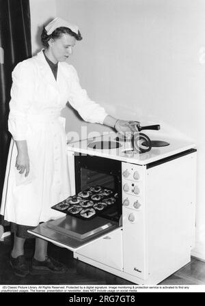 In cucina negli anni '1950 Una giovane donna in cucina al nuovissimo fornello elettrico Kockums. Viene mostrata la dimostrazione della funzione pratica di sollevare la piastra riscaldante per pulirla correttamente sotto e intorno ad essa. È visibile un piatto di pasticcini appena sfornati. Svezia 1957 Foto Stock