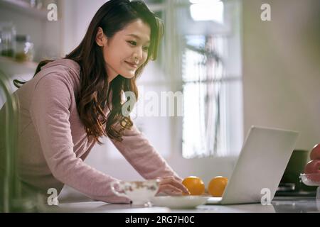 giovane donna d'affari asiatica che lavora in cucina a casa utilizzando un computer portatile Foto Stock