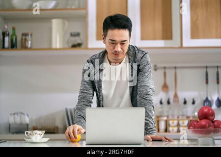 giovane uomo d'affari asiatico che lavora in cucina a casa usando un computer portatile Foto Stock