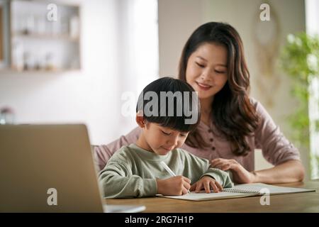 giovane madre asiatica seduta al tavolo a casa che aiuta il figlio con il suo studio Foto Stock