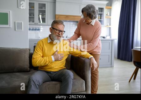 Uomo anziano che tocca il petto con attacco di cuore e sua moglie che lo sostiene Foto Stock