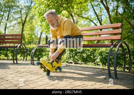 Uomo anziano dai capelli grigi che indossa pattini a rotelle mentre è seduto sulla panchina nel parco Foto Stock