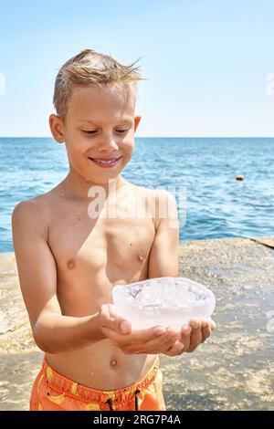 Il ragazzo in piedi sul molo di pietra tiene le meduse trasparenti con un'espressione divertente guarda le meduse in mano Foto Stock