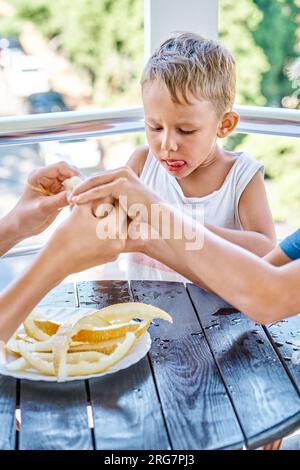 Il bambino in età prescolare mangia una fetta di melone fresco sul balcone dell'hotel. Il bambino biondo guarda le fette di frutta con un'espressione felice durante le vacanze estive Foto Stock