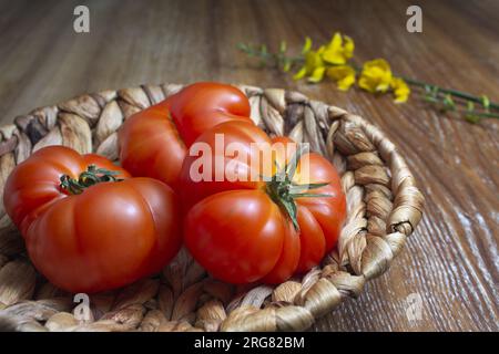 Primo piano della natura morta di tre pomodori freschi in un cestino. Atmosfera rustica in un tavolo di legno. Vista orizzontale. Foto Stock