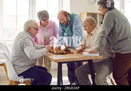 Gruppo di persone anziane felici e diverse che giocano a scacchi in casa di cura sedute a tavola. Foto Stock