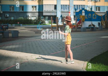 Una graziosa ragazza in età scolare soffia grandi bolle di sapone nel cortile di un edificio a più piani in estate. concetto estivo, giochi all'aperto Foto Stock