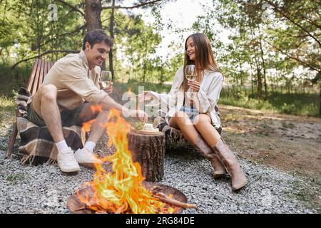 Coppia allegra con vino che parla seduti sulle sedie a sdraio vicino al cibo e alla legna da ardere all'aperto Foto Stock