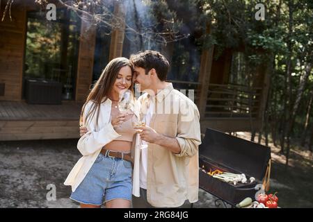 Uomo sorridente che abbraccia la fidanzata con del vino vicino a un barbecue sfocato e casa per le vacanze sullo sfondo Foto Stock