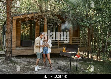 Coppia sorridente che brinda con vino e si trova vicino al barbecue e alla casa vacanze all'aperto Foto Stock