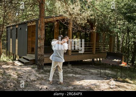 uomo felice che solleva una donna allegra con tatuaggio a portata di mano, casa vacanze nella foresta, vacanza estiva, amore Foto Stock