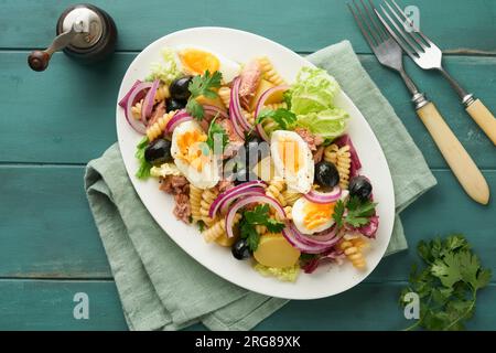 Insalata di tonno con pasta, uova, patate, olive, cipolle rosse e salsa in piatto bianco su fondo rustico in legno turchese. Insalata Nicoise. Fra Foto Stock