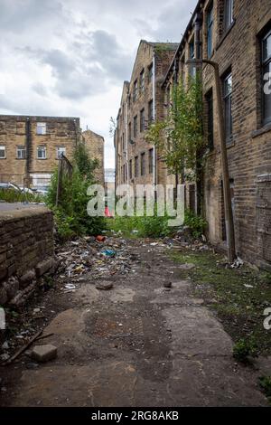 Vicolo desolato sulla strada posteriore dietro alcuni vecchi edifici di fabbrica/mulini a Bradford, Yorkshire, 2023. Mattoni e rifiuti sparsi lungo il percorso. Foto Stock