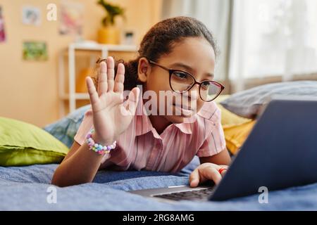 Intelligente studentessa afroamericana con gli occhiali che sventolano la mano e guardano lo schermo del notebook mentre si sdraiano sul letto e comunicano in videochat Foto Stock