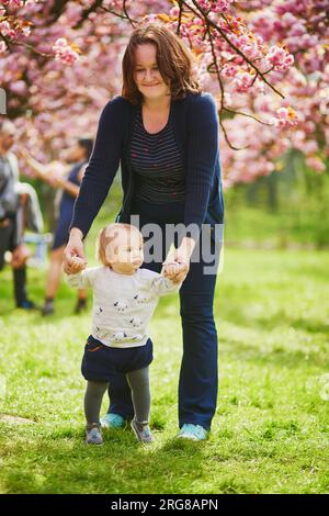 Giovane donna con bambina di 1 anno in parco in un giorno di primavera, che si gode la stagione della fioritura dei ciliegi. Madre che aiuta sua figlia a fare i primi passi. Bimbi piccoli l Foto Stock