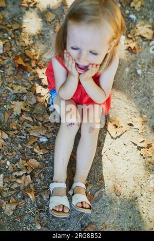 Una graziosa bambina seduta a terra che piange dopo essere caduta. Il bambino si fa male mentre cammina nel parco. Ragazzo che guarda il suo boo-boo Foto Stock