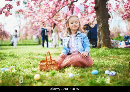 Bambina in età prescolare che indossa le orecchie del coniglio e gioca a caccia all'uovo a Pasqua. I bambini raccolgono uova colorate nel cestino. Bambino che festeggia la Pasqua all'aperto Foto Stock