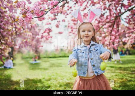 Bambina in età prescolare che indossa le orecchie del coniglio e gioca a caccia all'uovo a Pasqua. I bambini raccolgono uova colorate nel cestino. Bambino che festeggia la Pasqua all'aperto Foto Stock