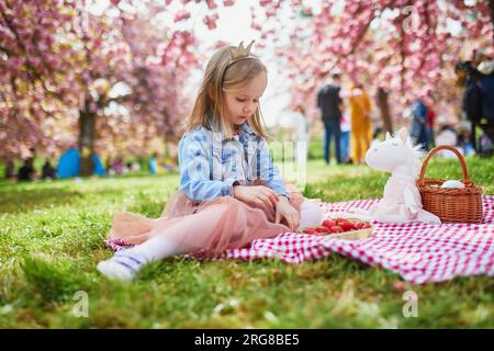 Adorabile bambina in età prescolare con gonna tutu e corona della principessa che si gode un picnic con unicorno giocattolo in una bella giornata primaverile nel giardino dei ciliegi in fiore Foto Stock