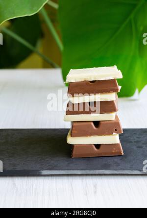 Quadrati bianchi, al latte e al cioccolato fondente impilati su un vassoio di ardesia con una foglia verde sullo sfondo. Shot verticale. Foto Stock