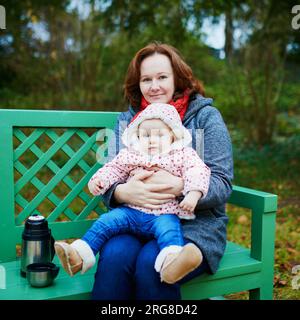 Madre e bambina che fanno un picnic nella foresta o nel parco, seduti sulla panchina con thermos Foto Stock