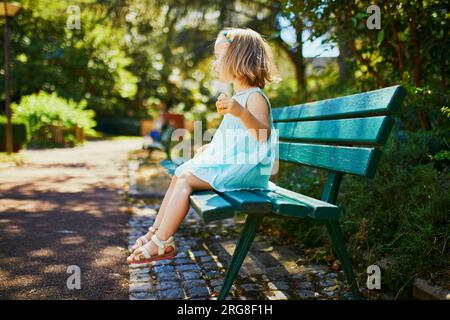 Adorabile bimba seduta sulla panchina all'aperto e che fa uno spuntino in una soleggiata giornata estiva. Un bambino felice che cammina nel parco. Passeggiate e alimentazione sana per Foto Stock