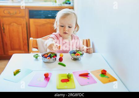 Adorabile bambina che gioca con frutta e verdura giocattolo a casa, all'asilo o all'asilo; cerca di abbinare figurina con l'immagine su una carta. Parole Foto Stock