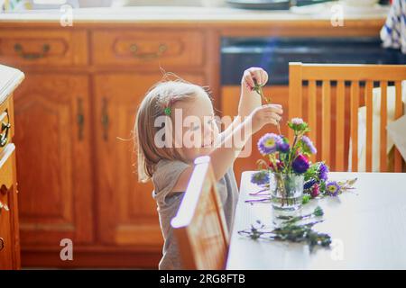 Adorabile bimba che prepara il bouquet di fiori a casa. Bambino che mette i pani in acqua. Attività domestiche per bambini Foto Stock