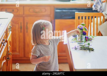 Adorabile bimba che prepara il bouquet di fiori a casa. Bambino che mette i pani in acqua. Attività domestiche per bambini Foto Stock