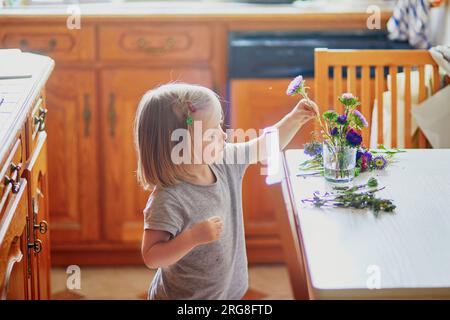 Adorabile bimba che prepara il bouquet di fiori a casa. Bambino che mette i pani in acqua. Attività domestiche per bambini Foto Stock