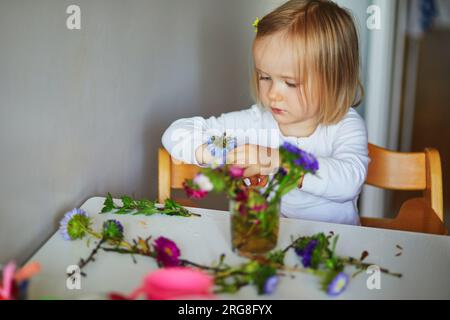 Adorabile bimba che prepara il bouquet di fiori a casa. Bambino che mette i pani in acqua. Attività domestiche per bambini Foto Stock